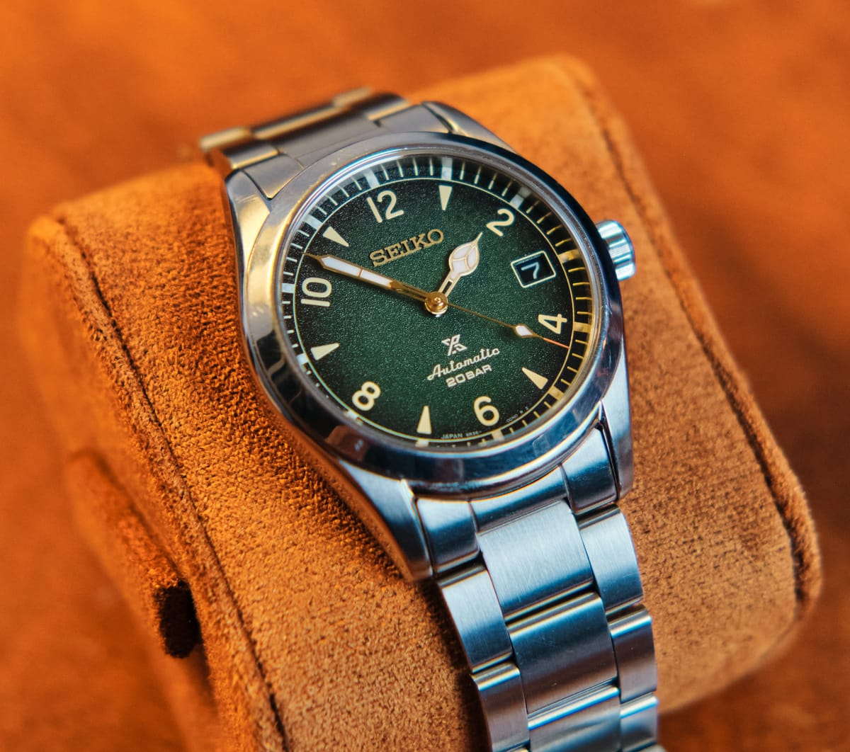 Close-up of a watch with a green dial on a steel bracelet, resting on a suede cushion