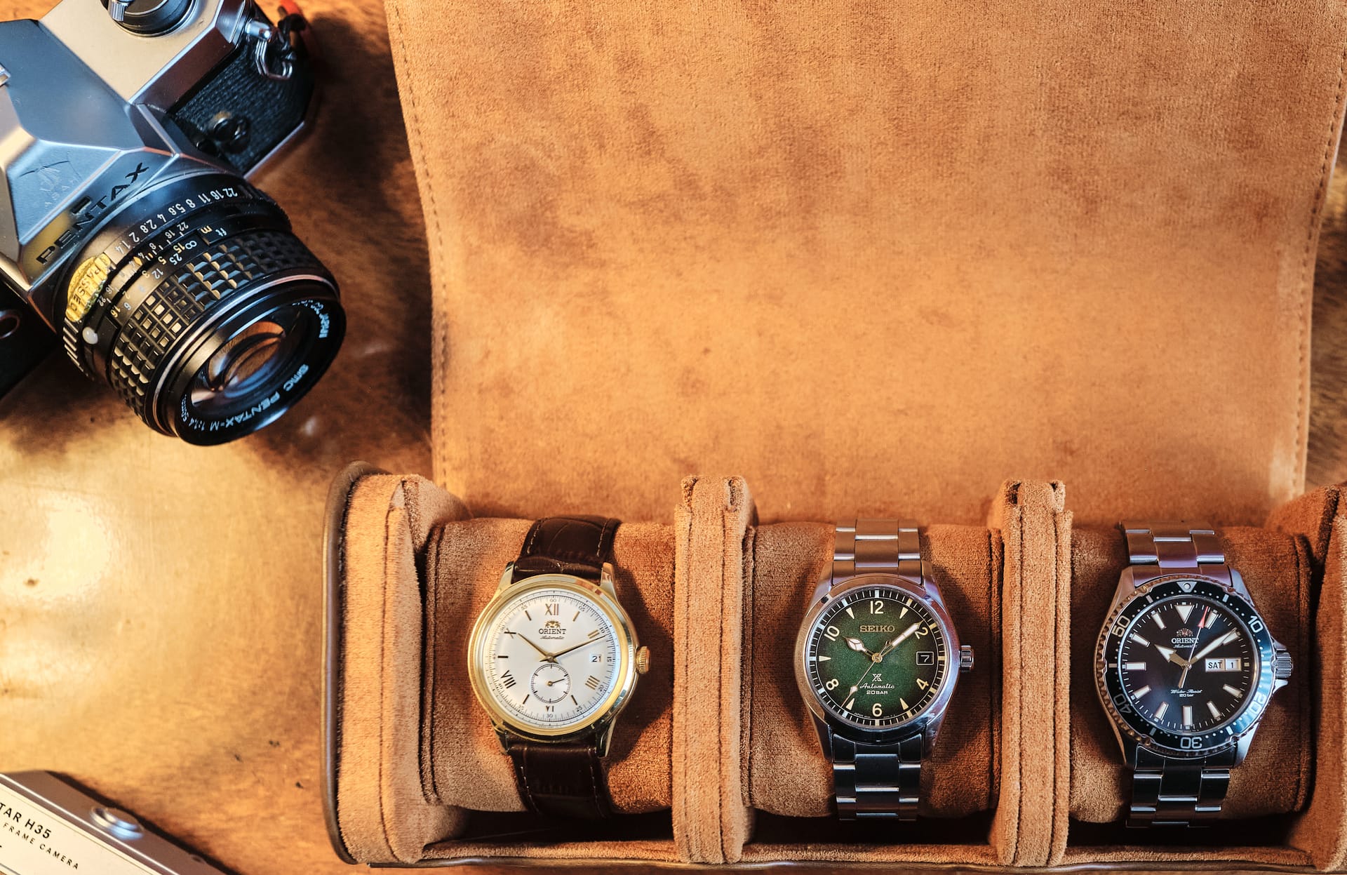 Three watches in an open suede watch roll on a wooden desk, beside two cameras